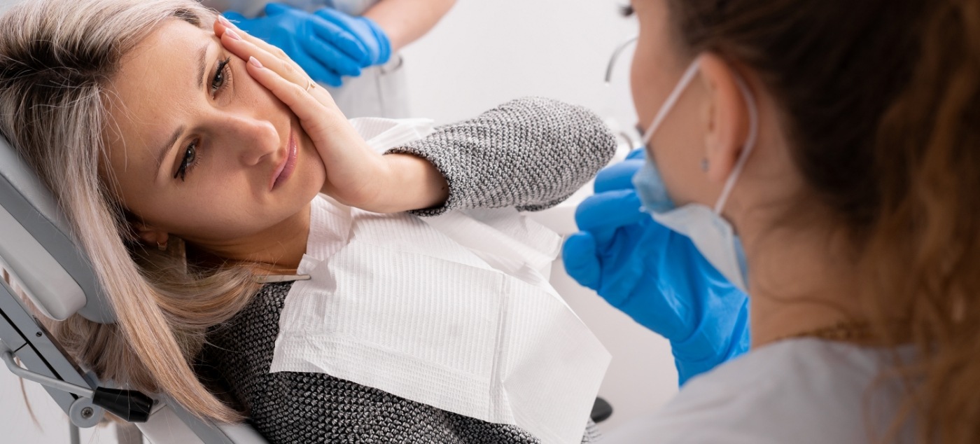 Wincing woman touching her cheek while talking to emergency dentist in Newark